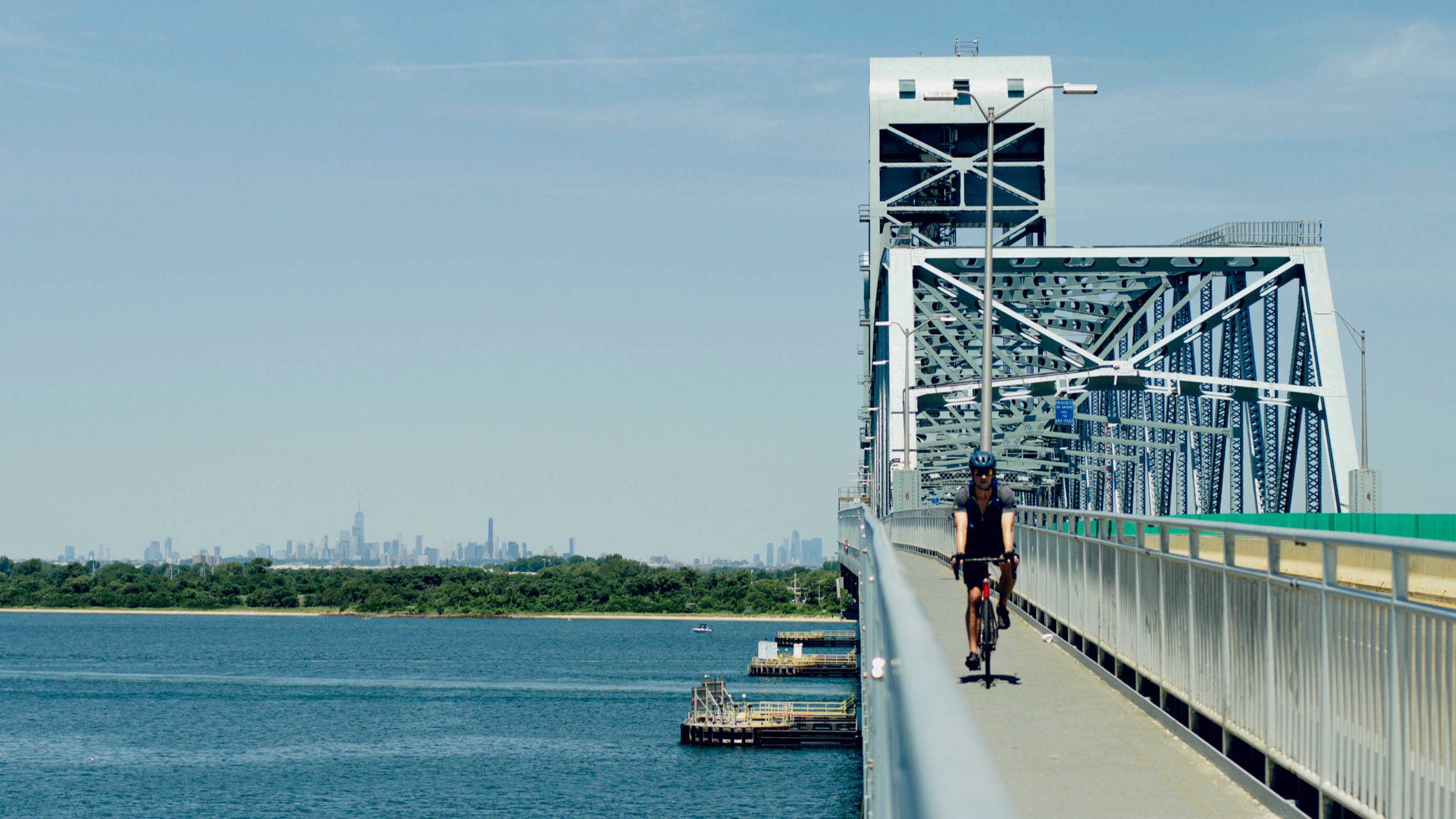 A person rides a bicycle on the pedestrian path of the Marine Parkway Bridge over blue water, with the Manhattan skyline in the distance.
