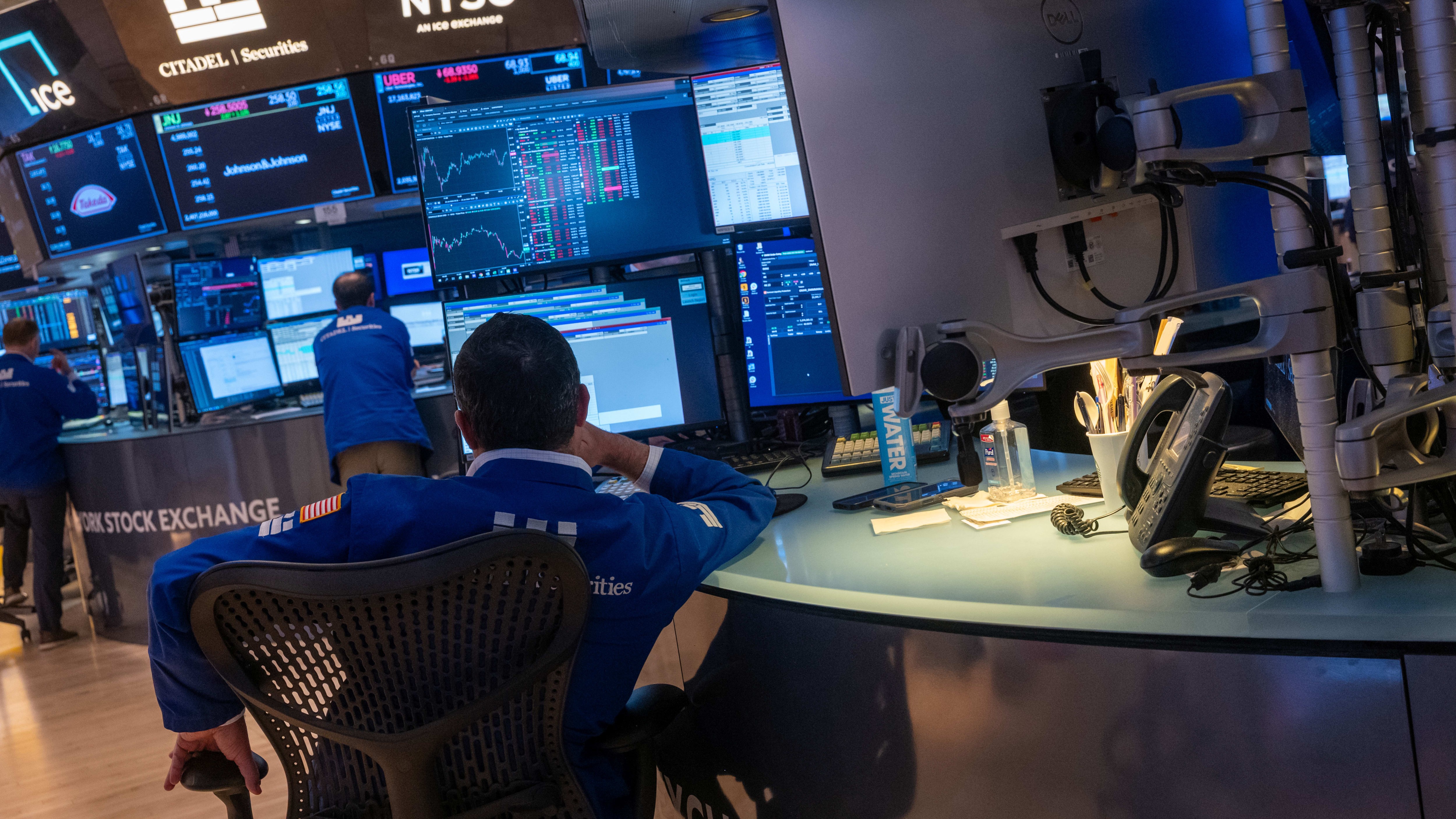 Traders in blue jackets work at computer stations on the floor of the New York Stock Exchange, with stock charts displayed on multiple monitors.