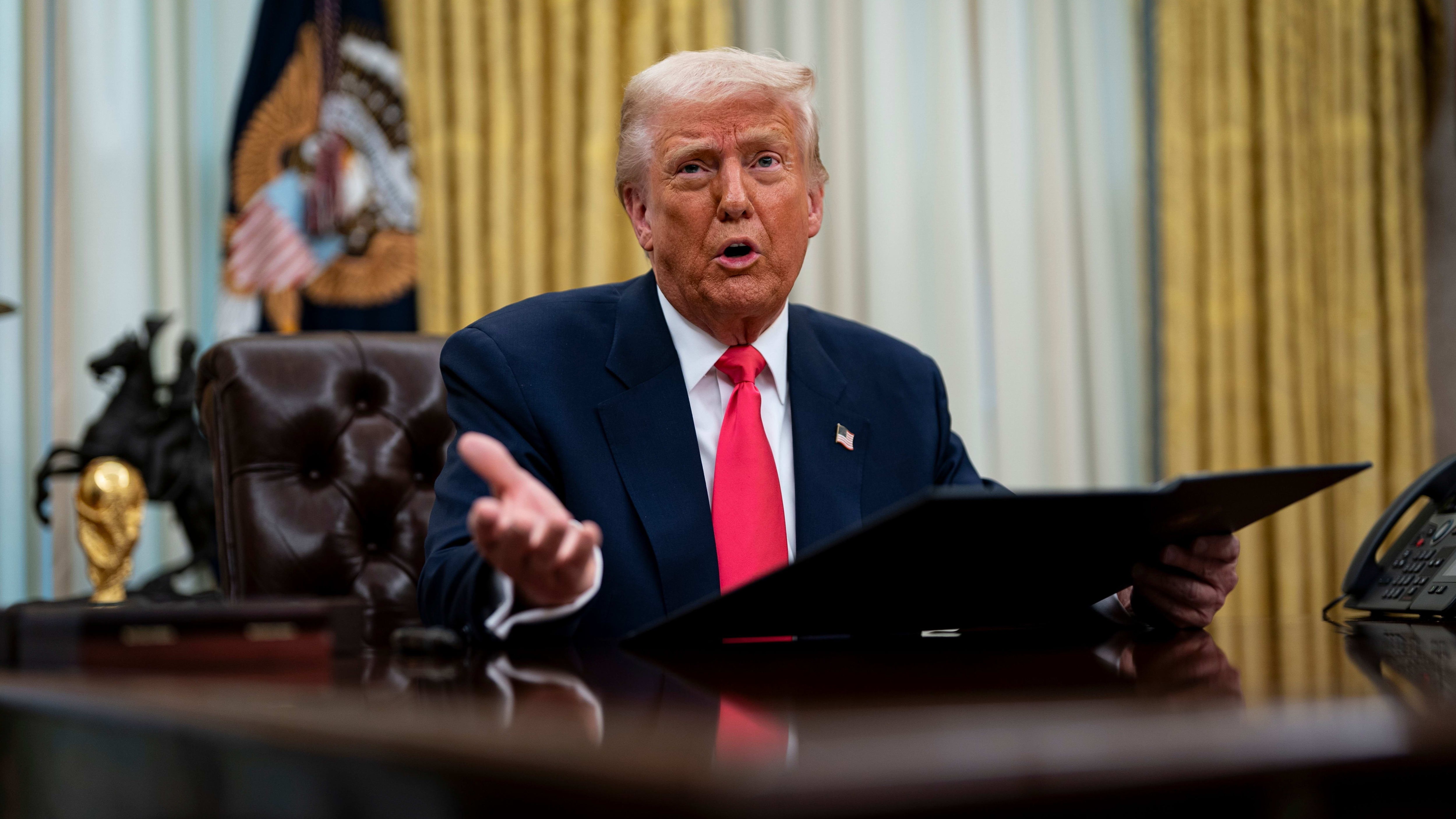 Donald Trump speaks while seated at a desk in the Oval Office, holding a folder.