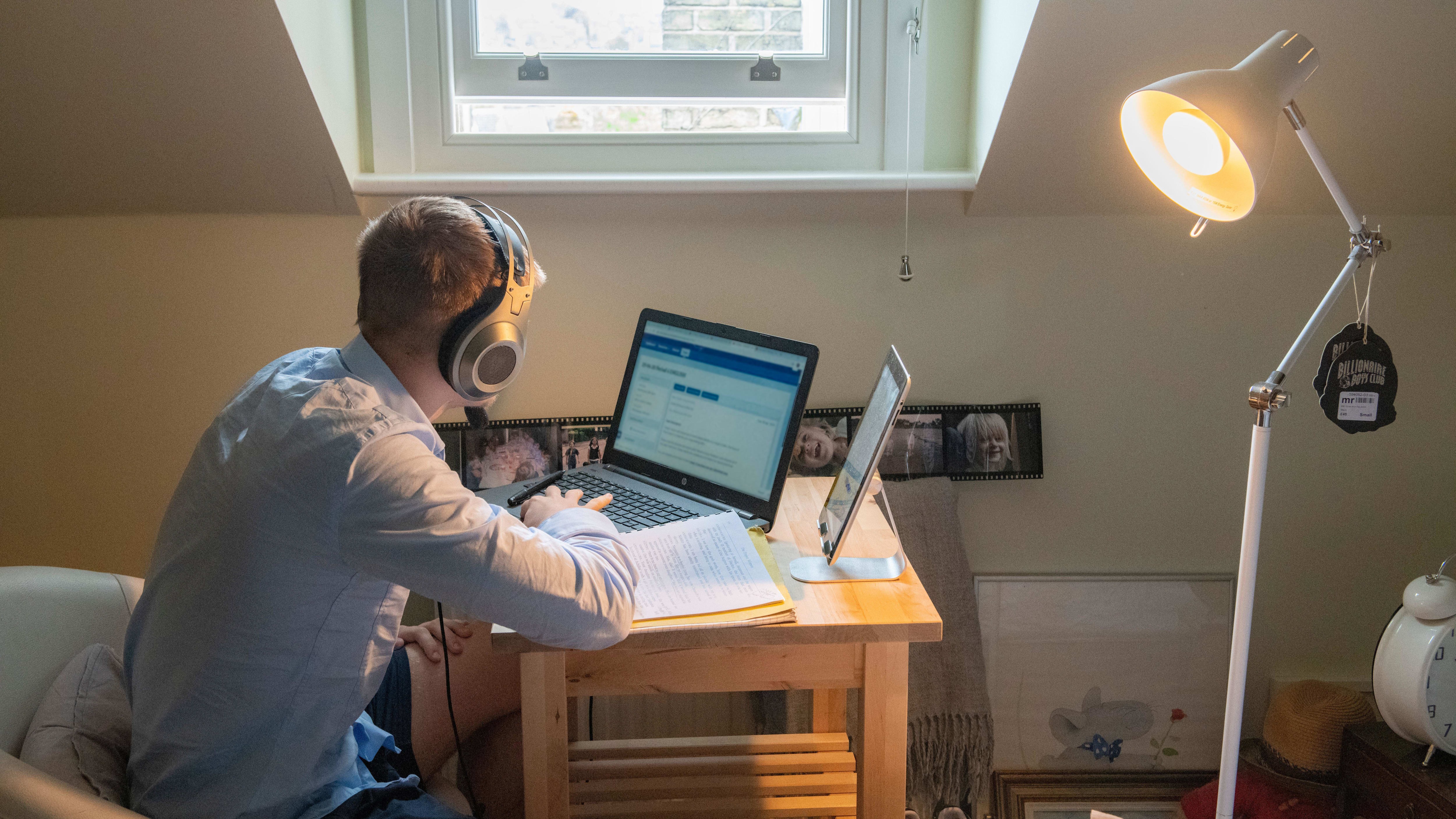 A teenage boy wearing headphones studies at a desk with a laptop and notebook during a remote school lesson.
