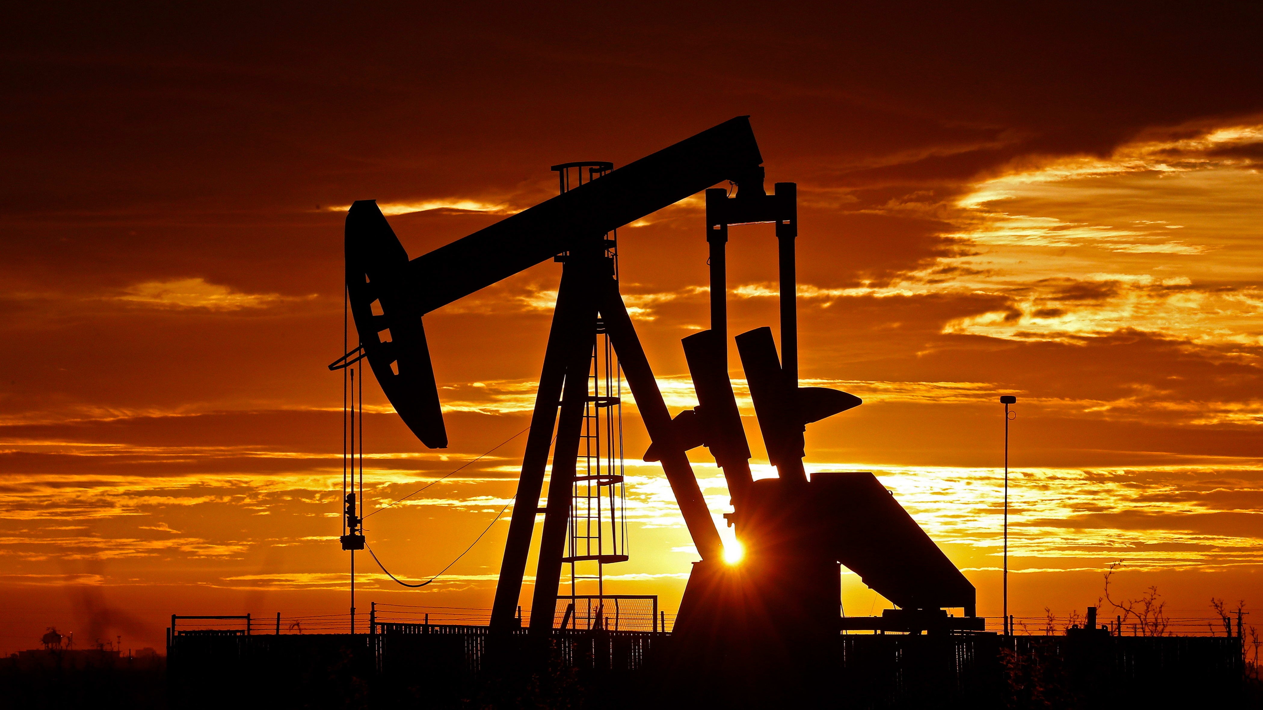 Pump jacks in silhouette operate in an oil field near Midland, Texas, with the sun rising and clouds lit orange behind them.