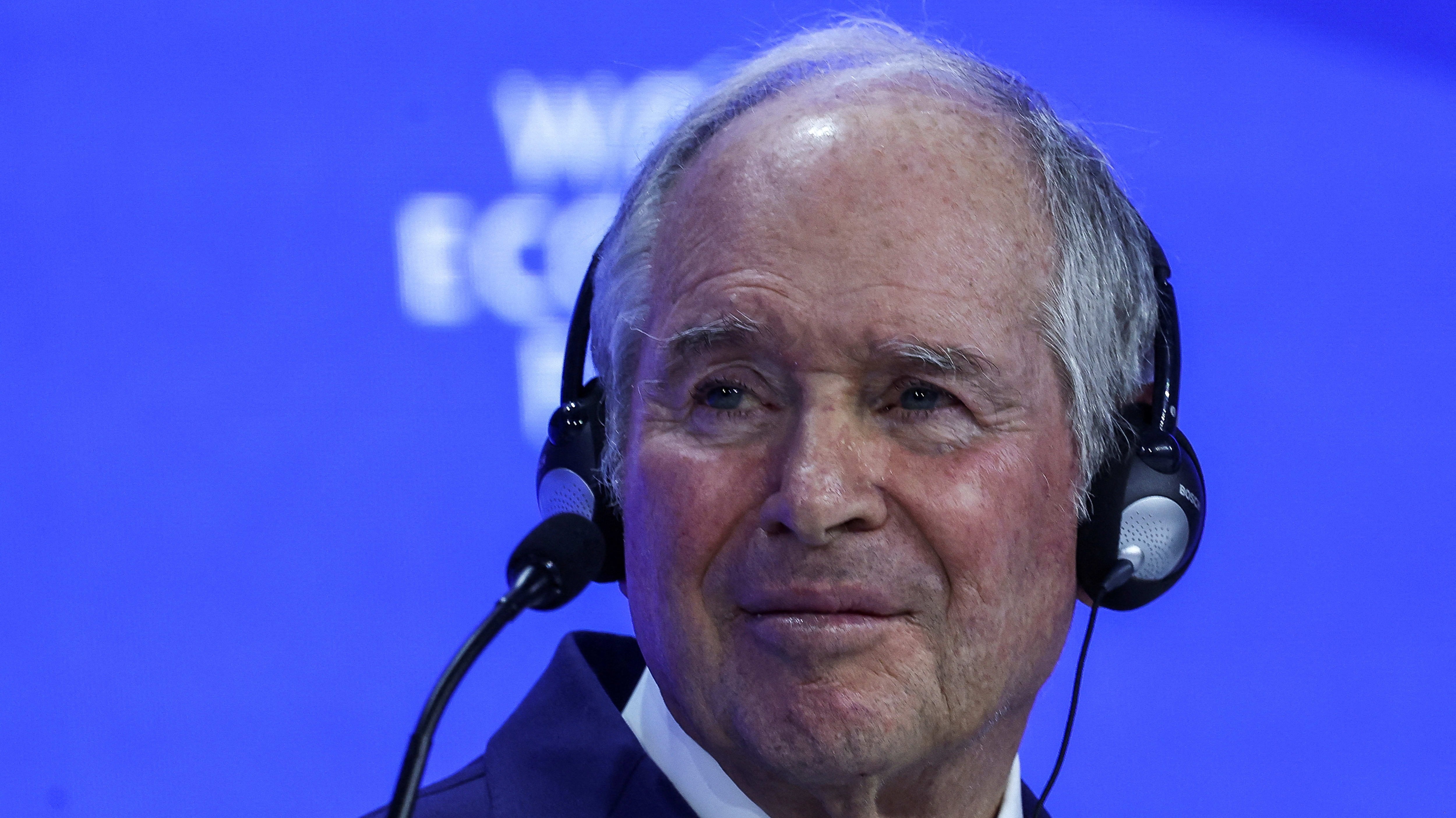 Stephen Schwarzman wearing a headset microphone, appearing to speak at the World Economic Forum with a blue background.