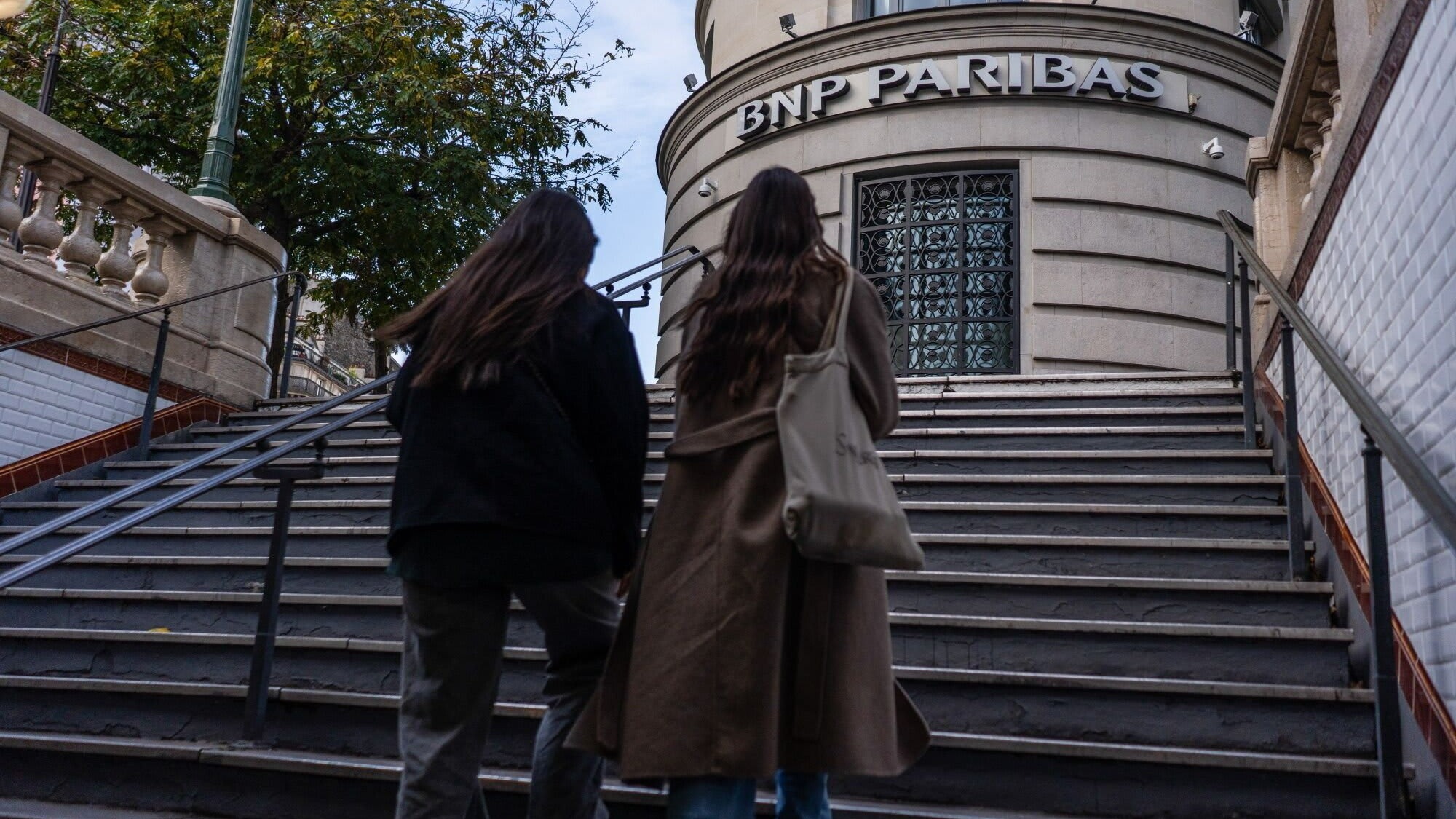 Two people walk up steps toward a BNP Paribas bank branch in Paris. The bank's sign is visible above the entrance.