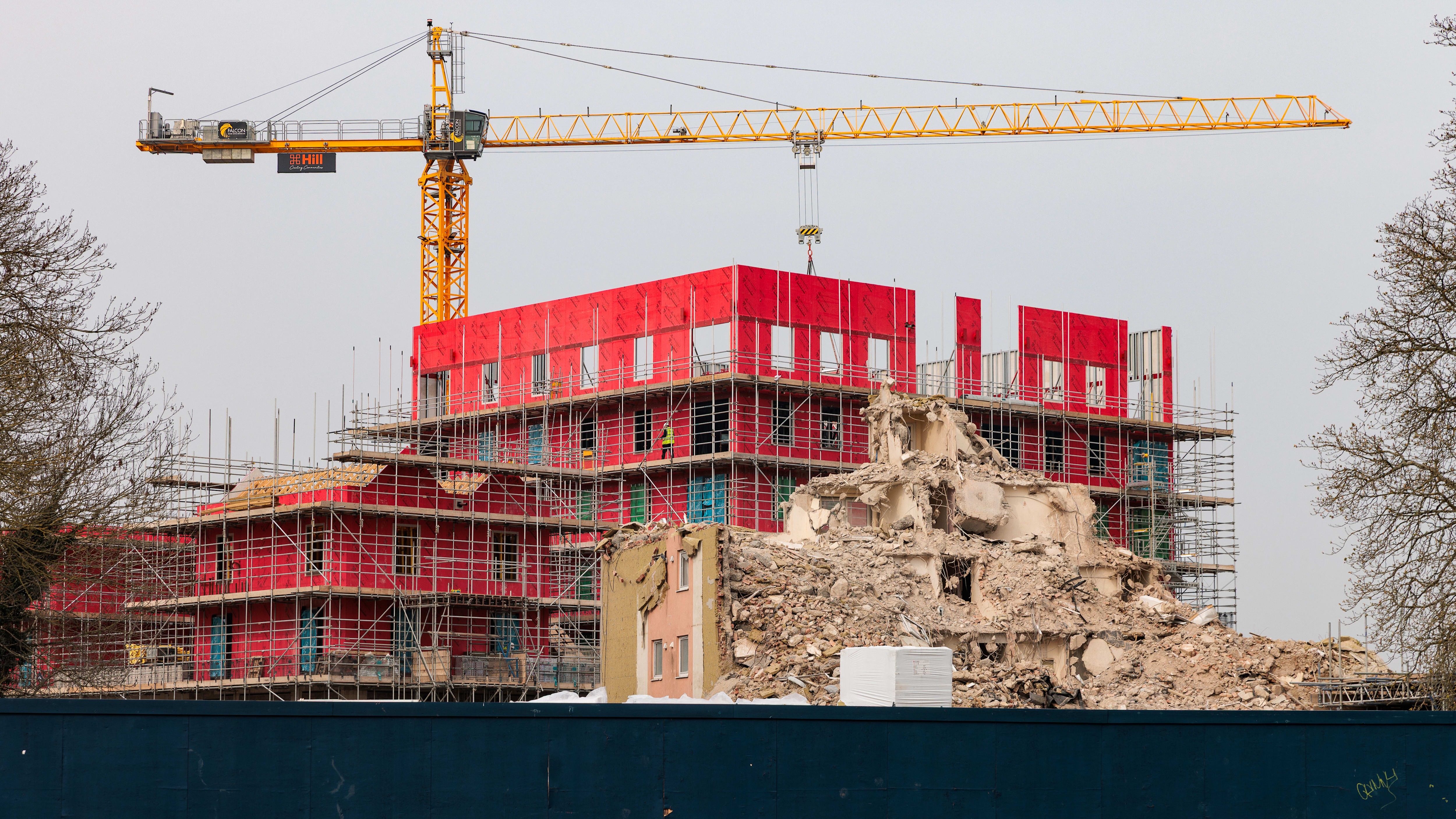 Partially demolished apartment block in front of a new building under construction, with scaffolding and a large yellow crane visible.