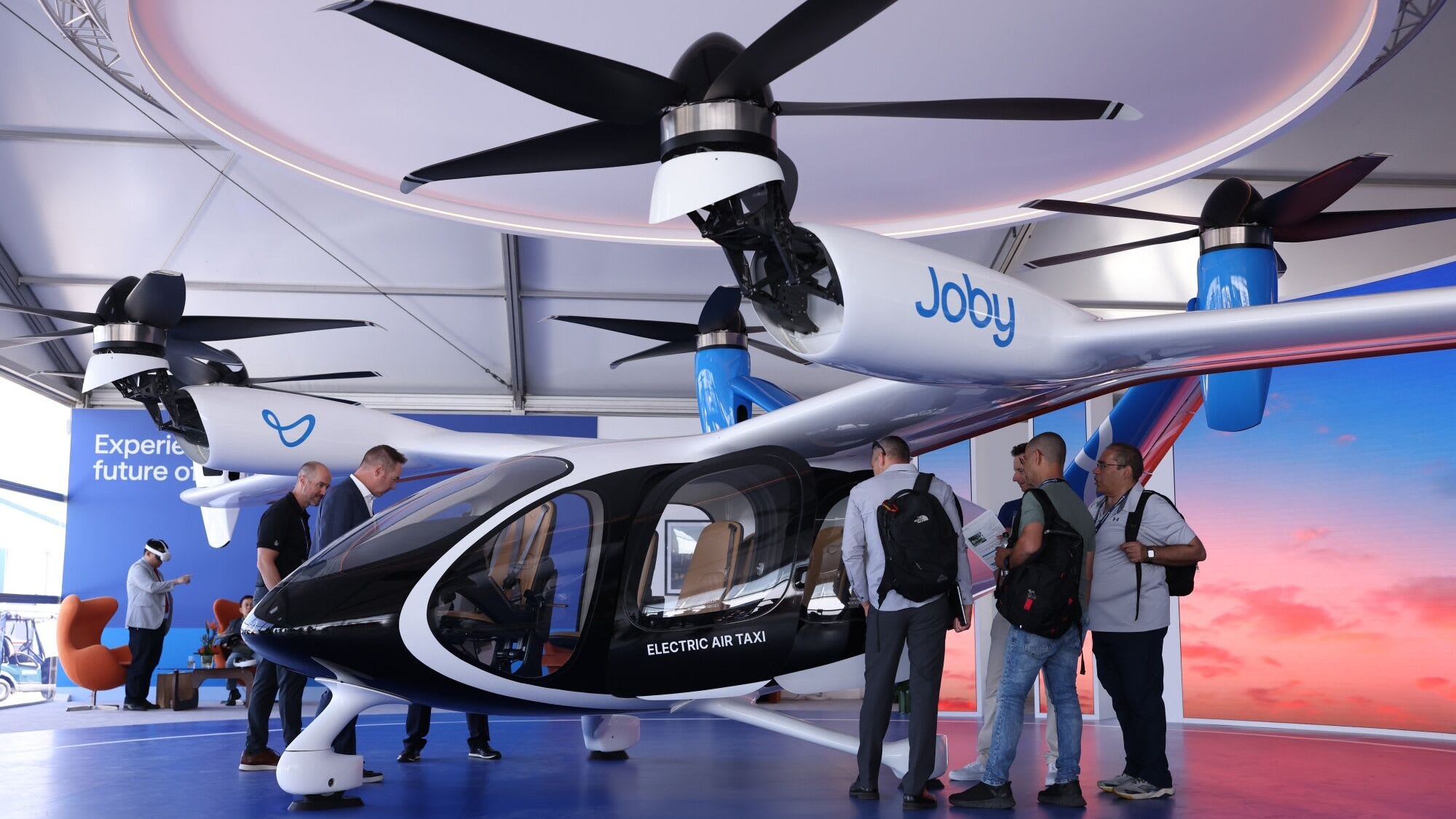 A Joby Aviation electric air taxi is displayed indoors as several people examine the aircraft at the Farnborough Airshow.
