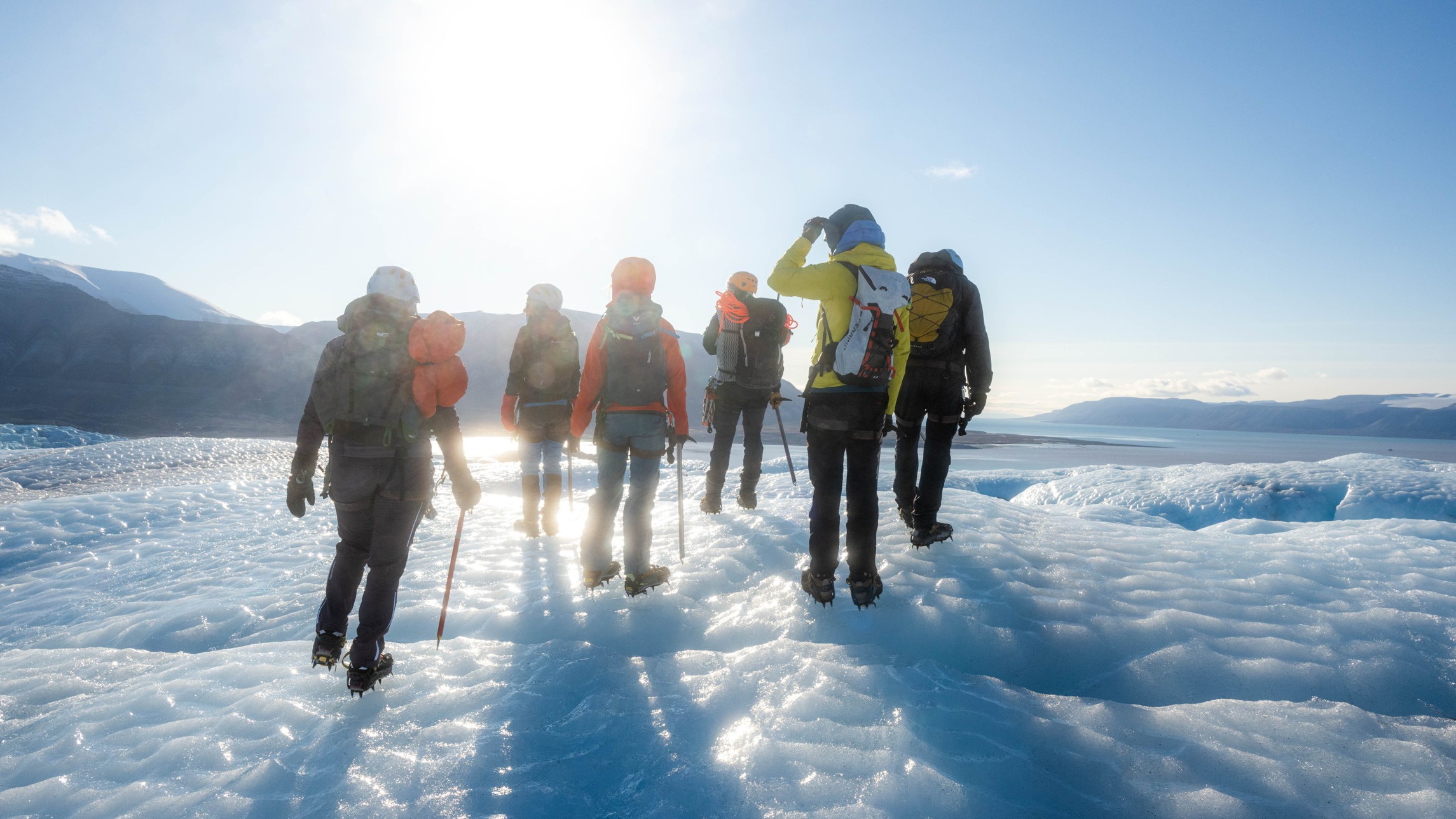 A group of people wearing winter gear and backpacks walk across an icy glacier under bright sunlight, with mountains and water visible in the distance.