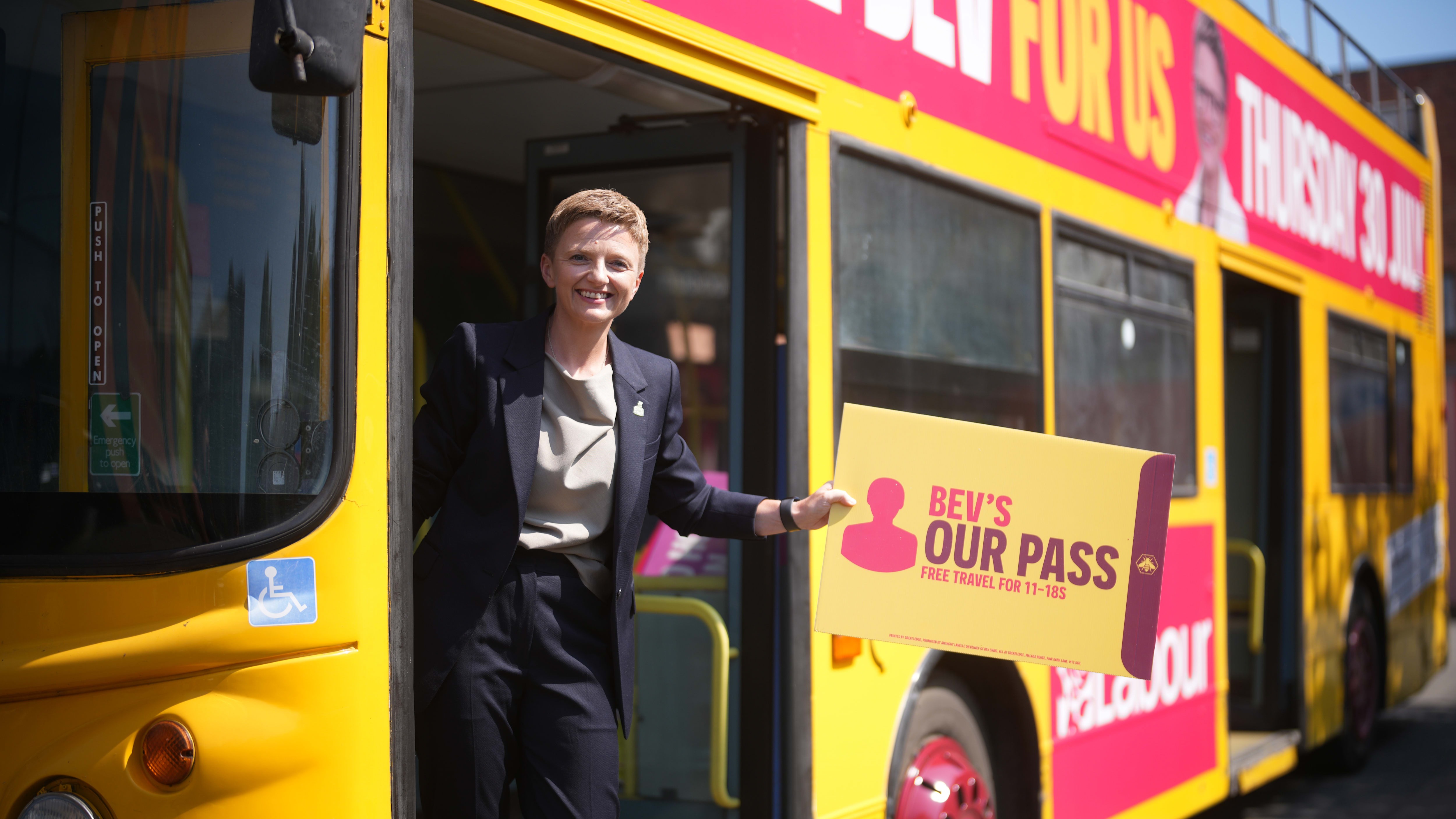 Bev Craig stands at the door of a yellow campaign bus, smiling and holding a sign promoting free travel for 11–18-year-olds.
