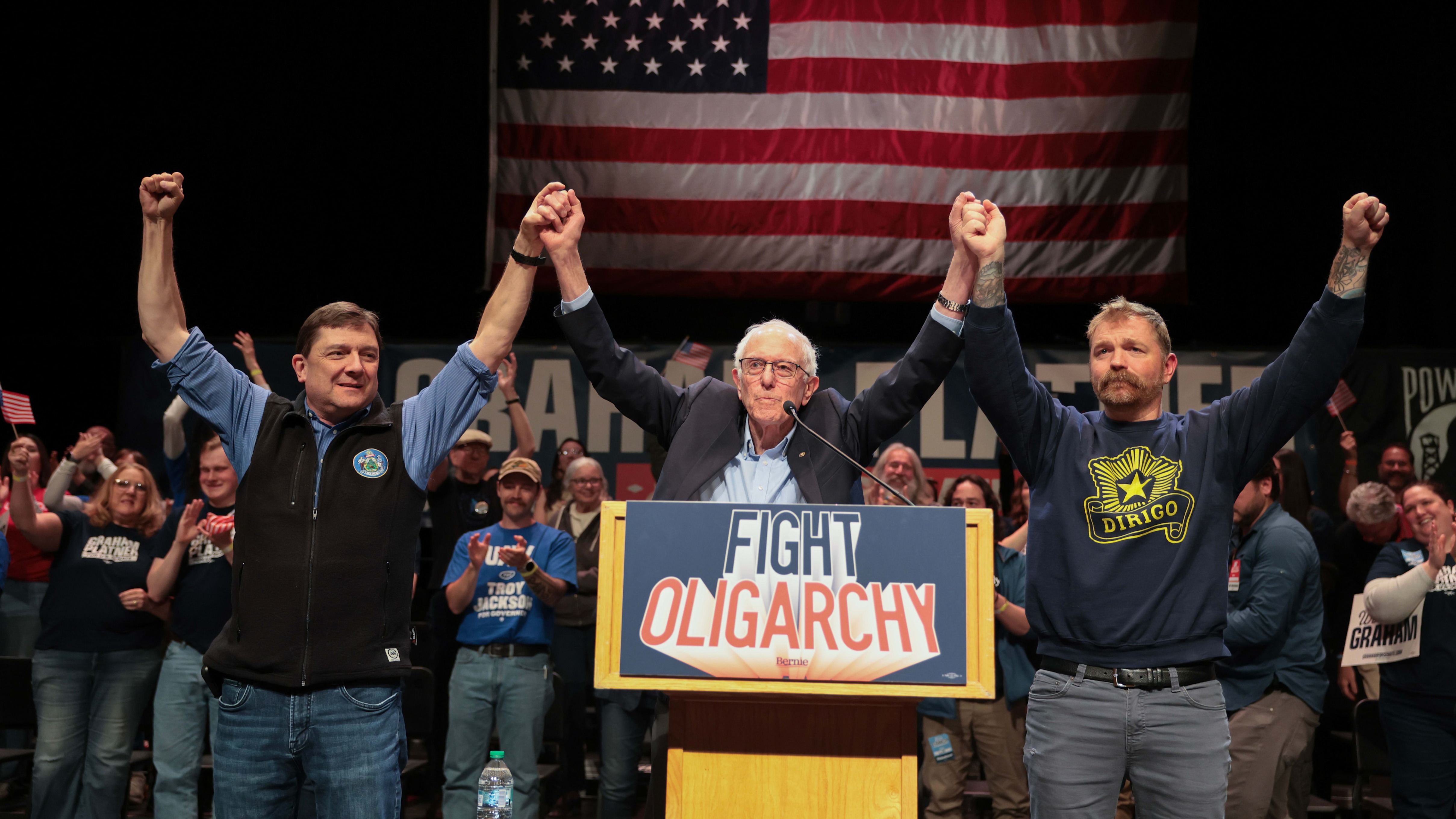 Sen. Bernie Sanders stands at a podium with Troy Jackson and Graham Platner, all raising joined hands before a cheering crowd.